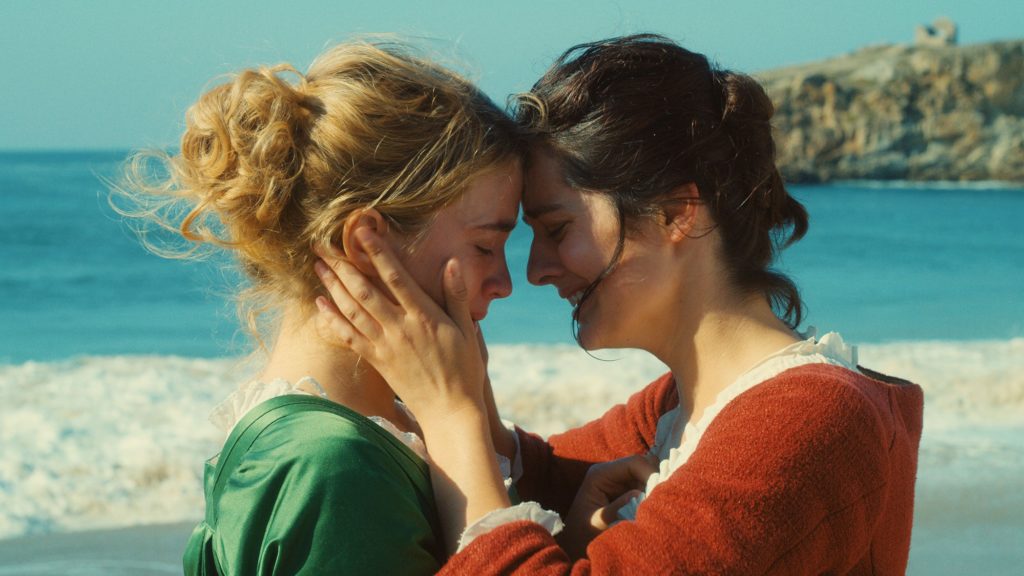 A photo still from the film Portrait of a Lady on Fire. The photo is a close up of Marianne and Héloïse with their foreheads touching, on the beach, with the ocean in the background.