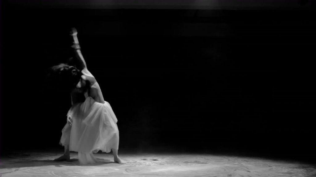 A black and white photo of dancer Shireen Talhouni, mid-performance, with an arm extended above her that is moving so fast it is blurred in the photo.
