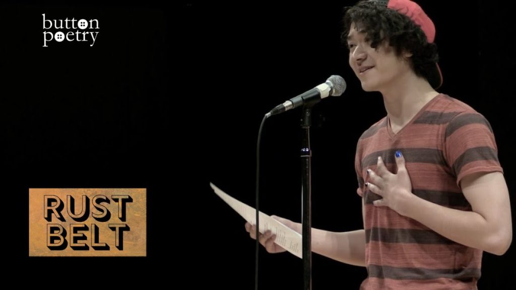 A photo of poet Spencer Brownstein mid-performance, standing onstage in front of a mic, holding a piece of paper in one hand and holding his other hand over his heart.