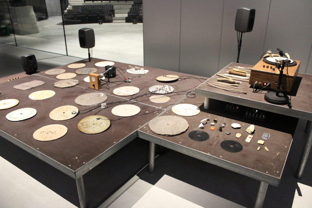 A photo of artwork The Ground by Tarek Atoui. The photo is of various instruments, plates, and sound systems on a raised platform.