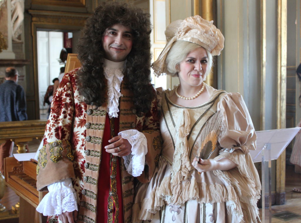 A photo of the opera singer and harpsichord player in the grand hall of the Château de Maisons. They are both wearing period piece costumes and wigs, and are smiling and posing for the camera.