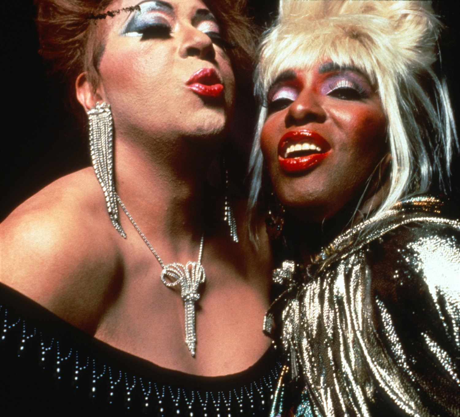 A close up photo of two ball performers from Paris is Burning, smiling and kissing towards the camera.