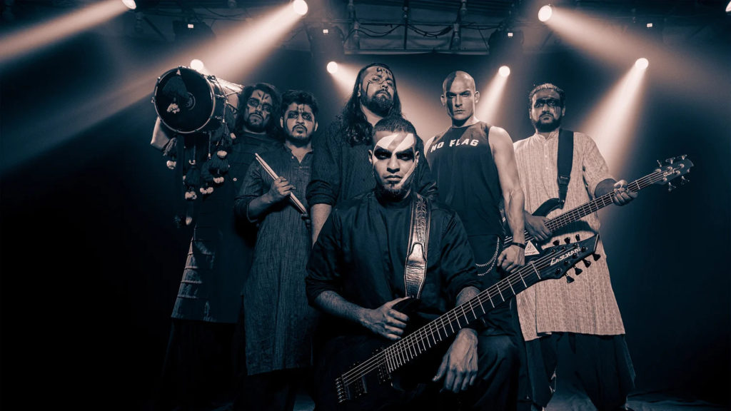 A promotional photo of Bloodywood. The members of the band are posed together, staring into the camera, holding their instruments and flanked by stage lighting.
