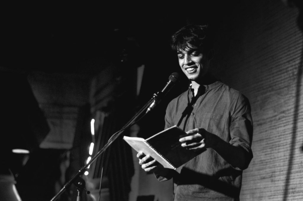 A photo of Rutwik Deshpande, standing on stage in front of a mic, reading from a book.