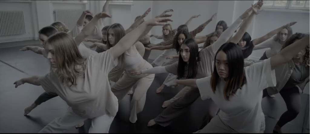 A photo still from the video by Unfolding Shapes. It shows several dancers, mid-performance, one arm raised in the arm, all dressed in grey.