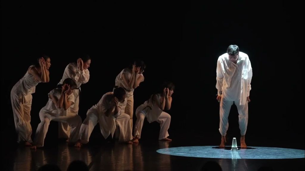 A photo still of a dance piece by Kim Jiwoo. It shows one dancer in white with a spotlight on him standing to the right, and a group of dancers in brown on the left all looking towards the dancer in white.