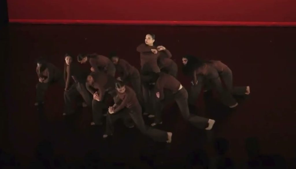 A photo of a dozen dancers onstage, mid-performance. All but one are hunched over, looking at the ground. They are all in black against a red backdrop.