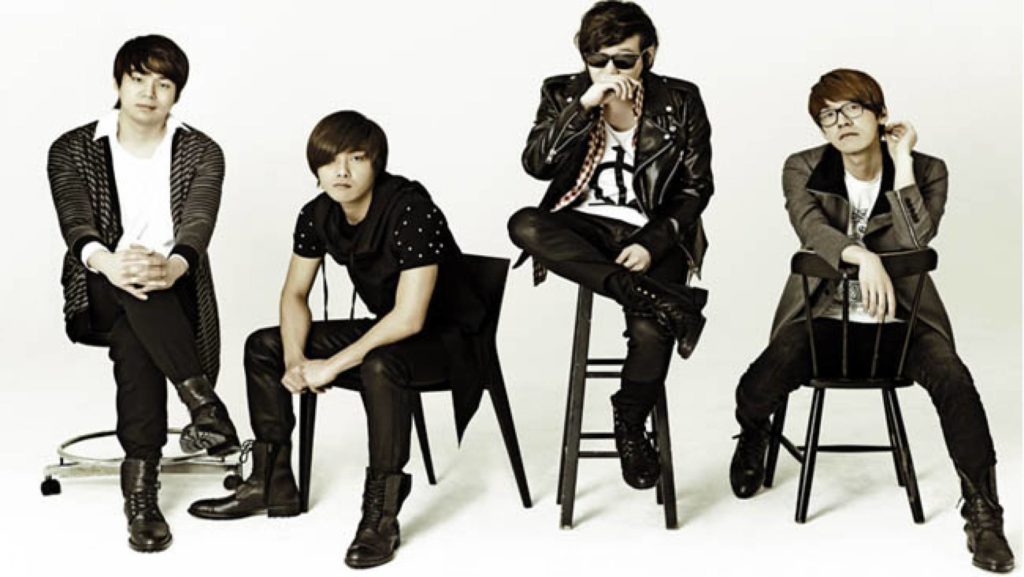 A promotional photo of Nell. The four band members are wearing black and white outfits, sitting on stools in a line, against a white backdrop.