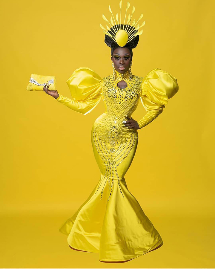A photo of a fashion design by Diego Montoya. The model is wearing a yellow dress and headpiece, and is standing against a yellow backdrop.
