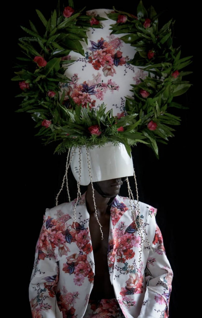 A photograph by Chris Schwagga. It depicts a Black man wearing a white and pink floral suit jacket and a white, pink, and green floral headpiece that covers the top half of his face.